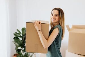Side view smiling lady in casual green shirt carrying cardboard box while preparing for moving in new home and looking at camera happily