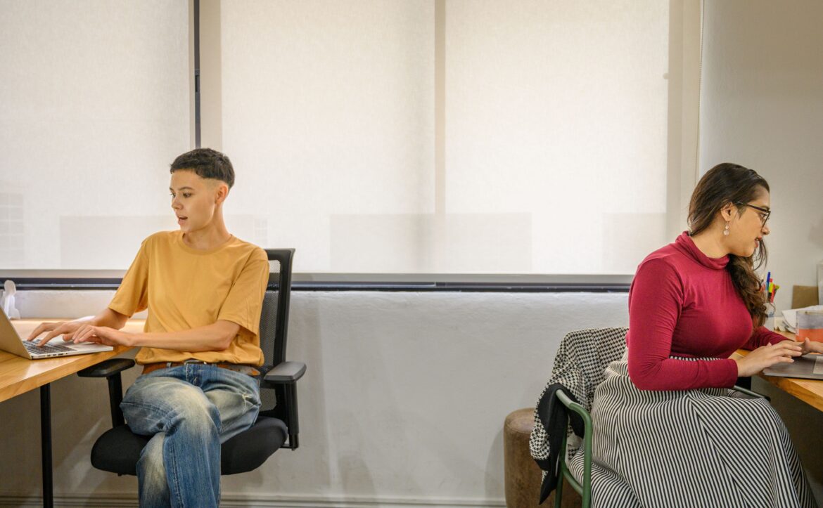Two coworkers sit at separate desks in a bright office with large window shades and natural light.