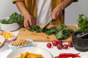 A person chops fresh kale on a wooden cutting board beside bowls of vegetables and other meal ingredients.