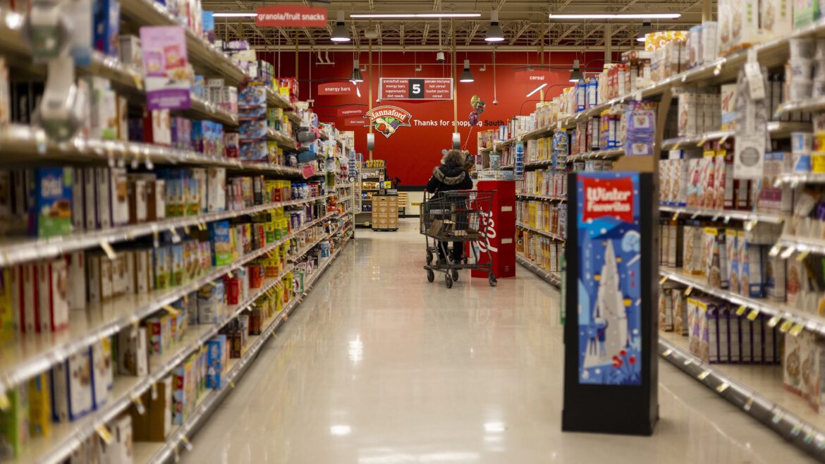 A shopper pushes a cart down a brightly lit supermarket aisle lined with densely stocked shelves.