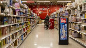 A shopper pushes a cart down a brightly lit supermarket aisle lined with densely stocked shelves.
