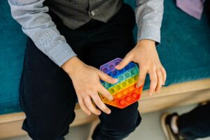 A child's hands press a rainbow pop-it fidget toy while seated on a teal bench.